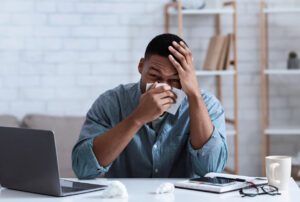 Ill Black Man Blowing Nose In Napkin Sitting In Office